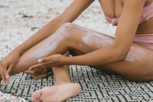 A tanned woman on the beach applying a thick layer of sunscreen to her legs.