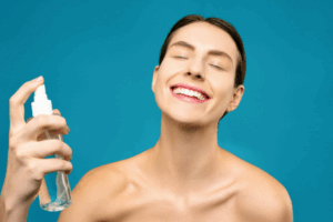 A young, smiling woman with glowing skin, spritzing a skincare product on her face.