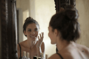 A young woman smiling and applying skincare in the mirror.