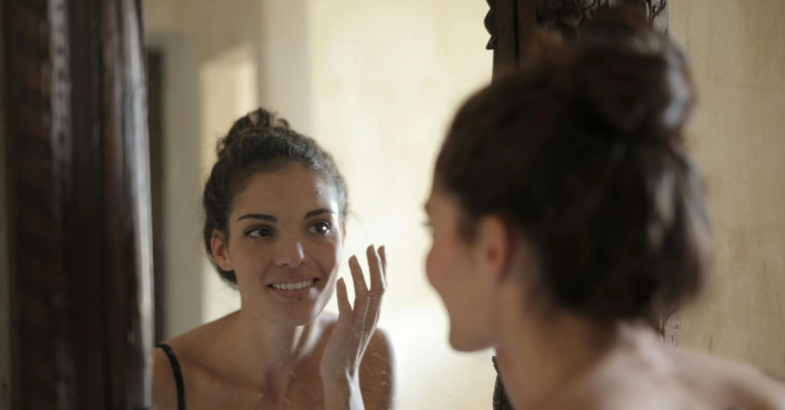 A young woman smiling and applying skincare in the mirror.