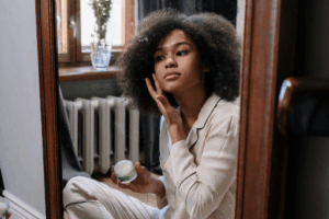 A young woman looking in a full-length mirror, applying a face cream.