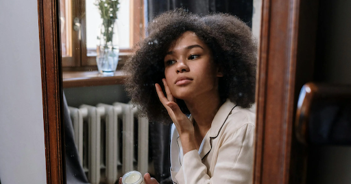 A young woman looking in a full-length mirror, applying a face cream.
